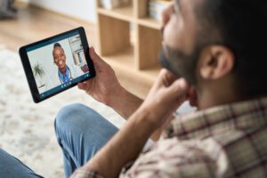 Young indian latin bearded businessman having videocall meeting at home with black female healthcare doctor therapist using tablet computer pointing to throat. Online virtual telemedicine health care concept.