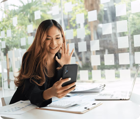 Woman waving and smiling during a video call on her phone, with a laptop open on the desk beside her — joining a Phone.com video meeting from a mobile device