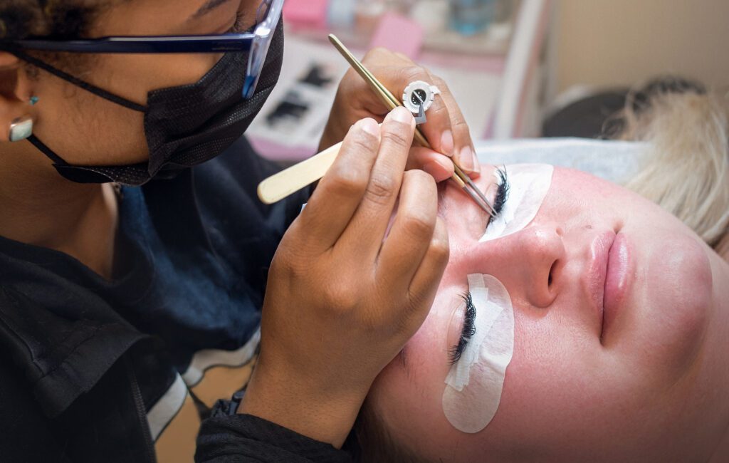 In a professional beauty salon specializing in facials, skincare, massage, lashes, and brows, a 26-year-old Latina lash technician applies eyelash extensions to a 31-year-old Caucasian blonde client. The image shows a detailed close-up of the precise lash application process with under-eye pads and tools, highlighting expert care and beauty services.